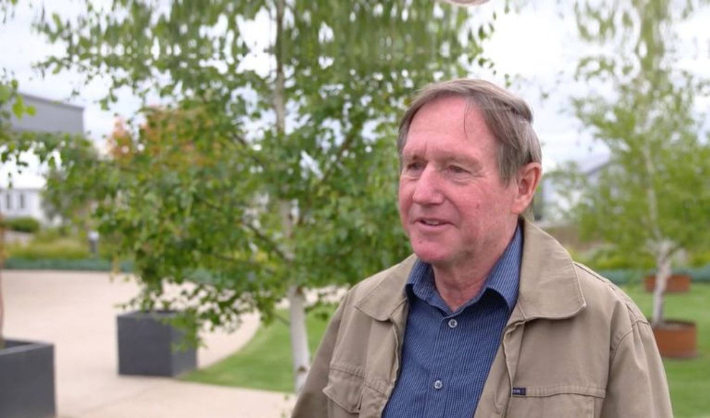 A man in  anavy shirt and tan jacket talks to the camera.
