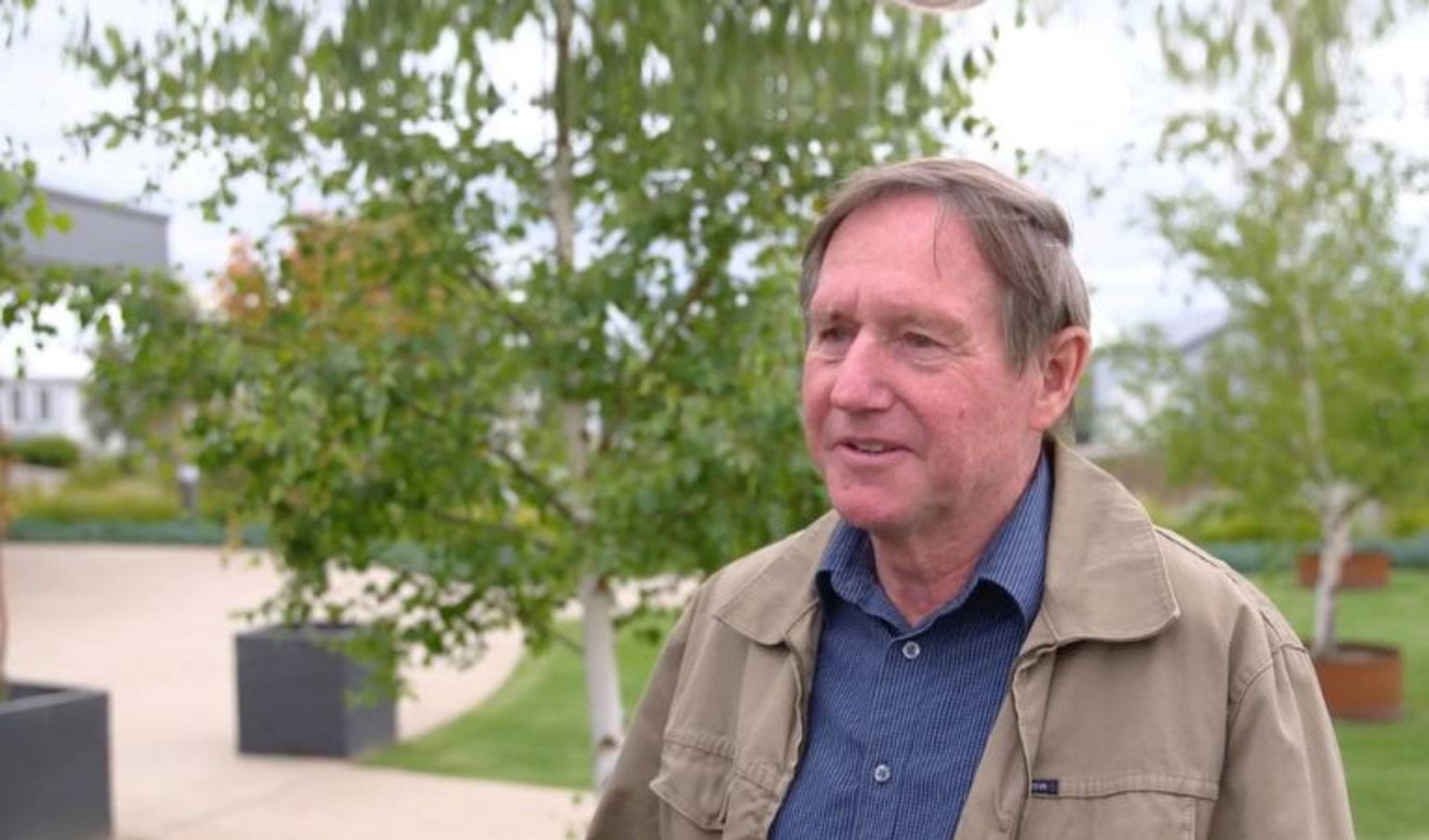 A man in  anavy shirt and tan jacket talks to the camera.