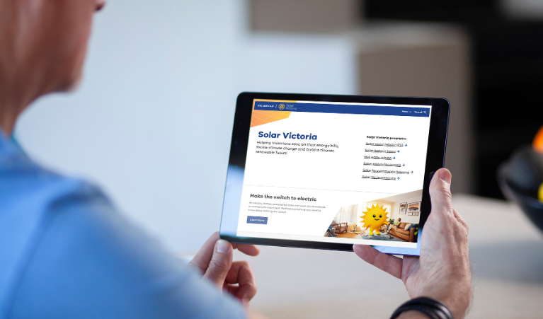 Person holding a tablet displaying the Solar Victoria website, showing information about solar rebates and switching to electric energy, with a clean modern interface and a home interior in the background.