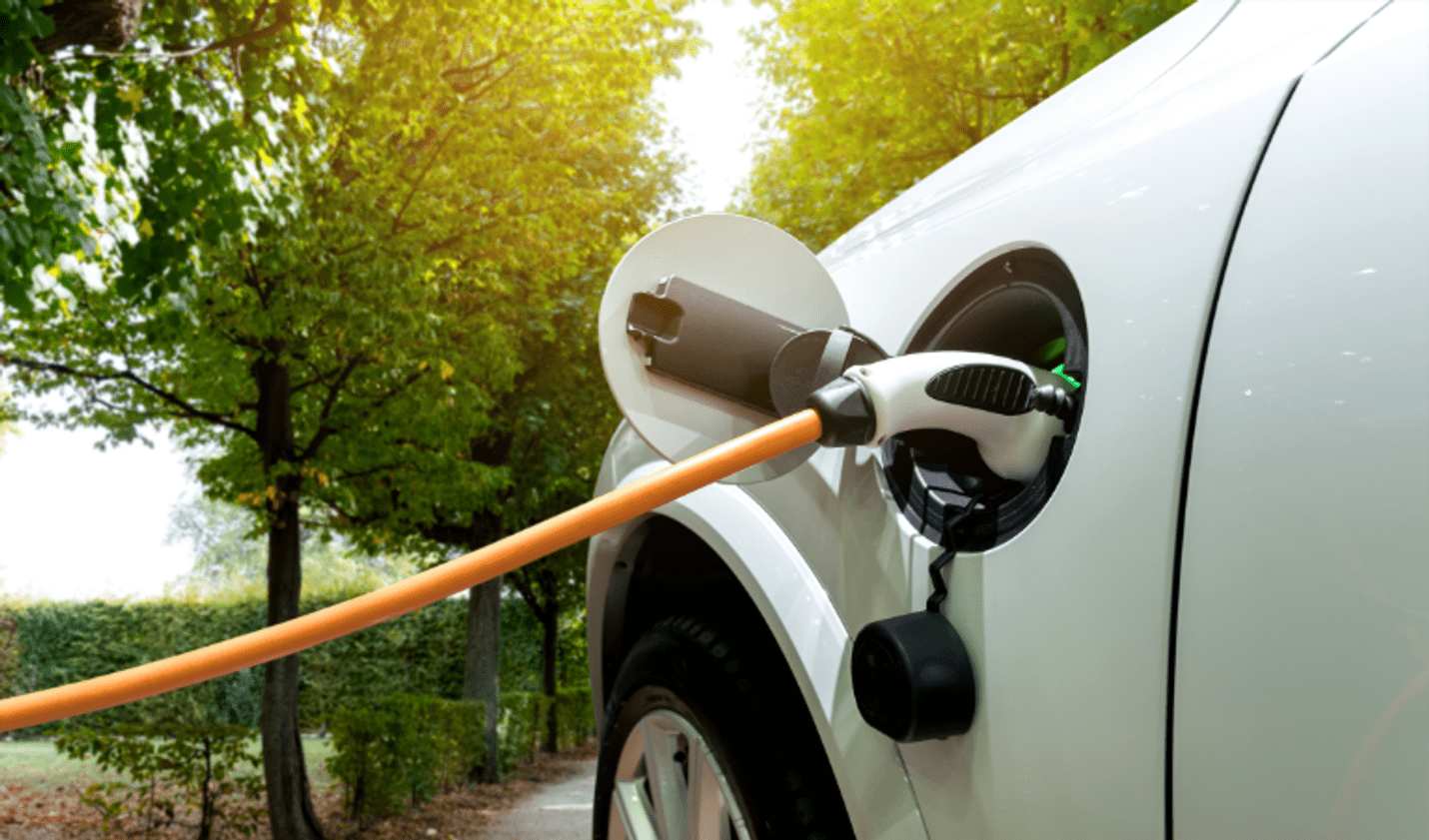 Close-up of an electric vehicle charging at a home EV charging station, with a charging cable connected to the vehicle in a leafy outdoor setting, highlighting clean energy transport and electric vehicle adoption.