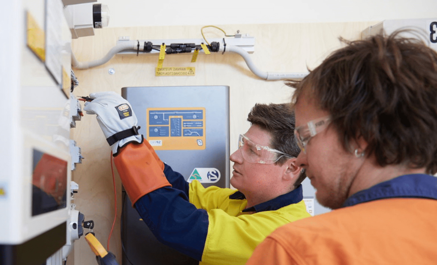 Trainees wearing safety glasses and protective gloves perform electrical testing and equipment maintenance on a switchboard during a hands-on electrification skills training program, demonstrating practical electrical industry skills in a technical workshop setting.