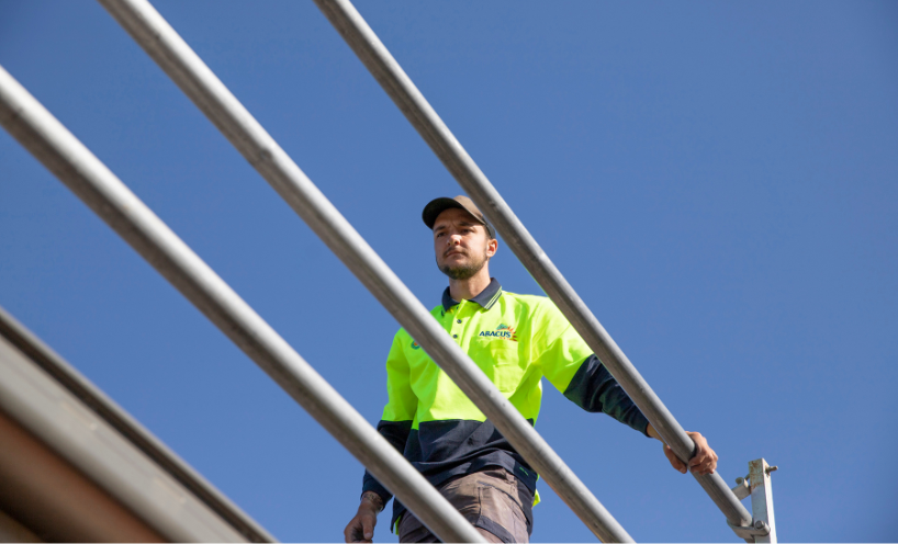 Crackdown on falls risks in the solar sector. A worker in high-visibility safety clothing stands on an elevated structure behind metal guardrails, viewed from below against a clear blue sky, highlighting the importance of fall prevention and safe working practices when working at height.