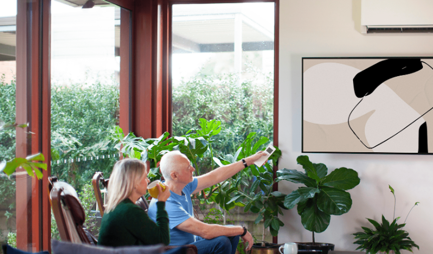 Two people relax in a living room while using a remote control to operate a wall-mounted split-system air conditioner in an all-electric home.