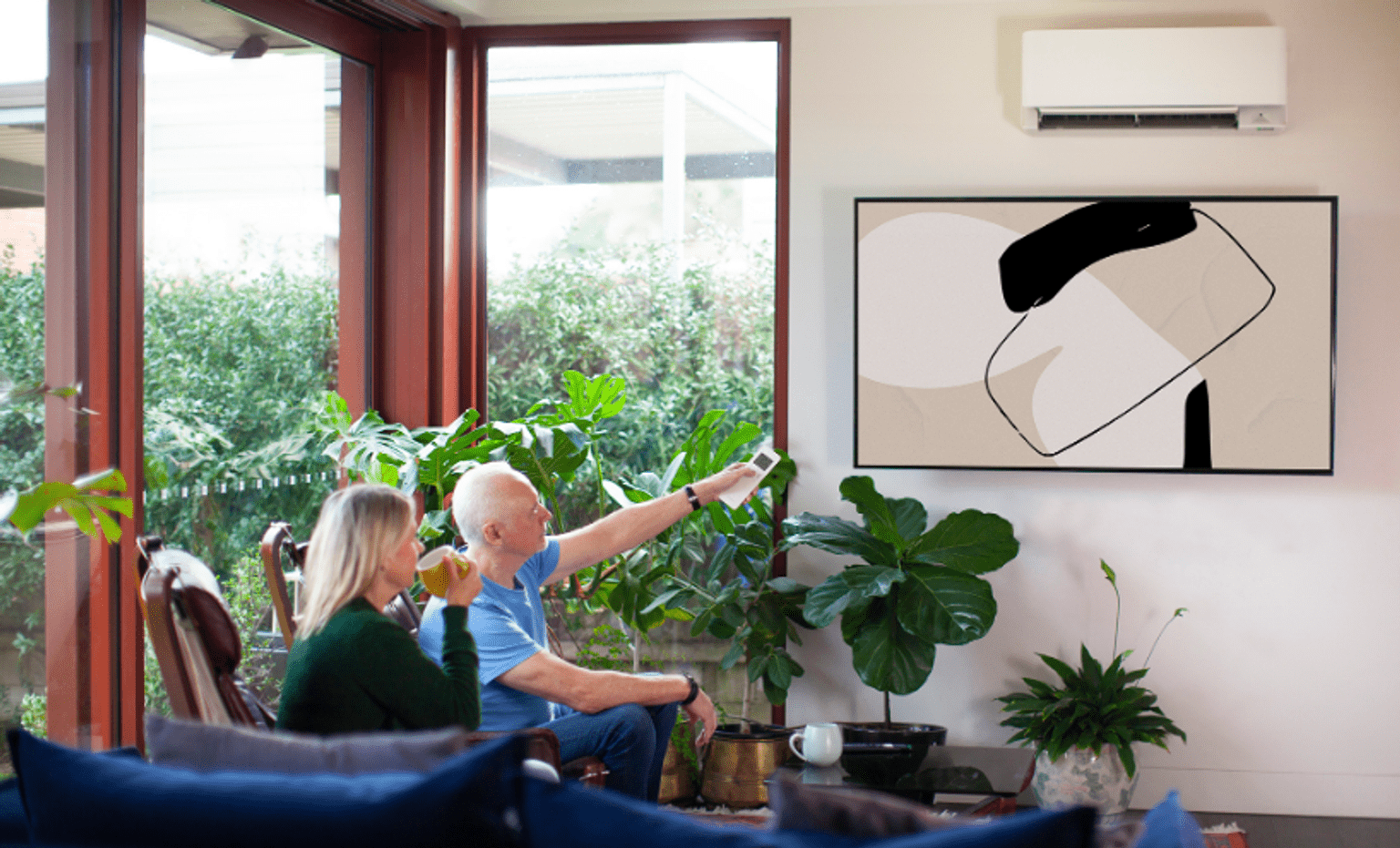Two people relax in a living room while using a remote control to operate a wall-mounted split-system air conditioner in an all-electric home.